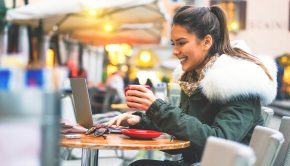 Beautiful and smiling young girl surfing on internet on her laptop while drinking a coffee - Stunning woman sitting outside in a cafe bar shopping online on her computer tablet booking holidays