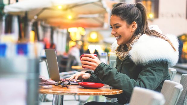 Beautiful and smiling young girl surfing on internet on her laptop while drinking a coffee - Stunning woman sitting outside in a cafe bar shopping online on her computer tablet booking holidays